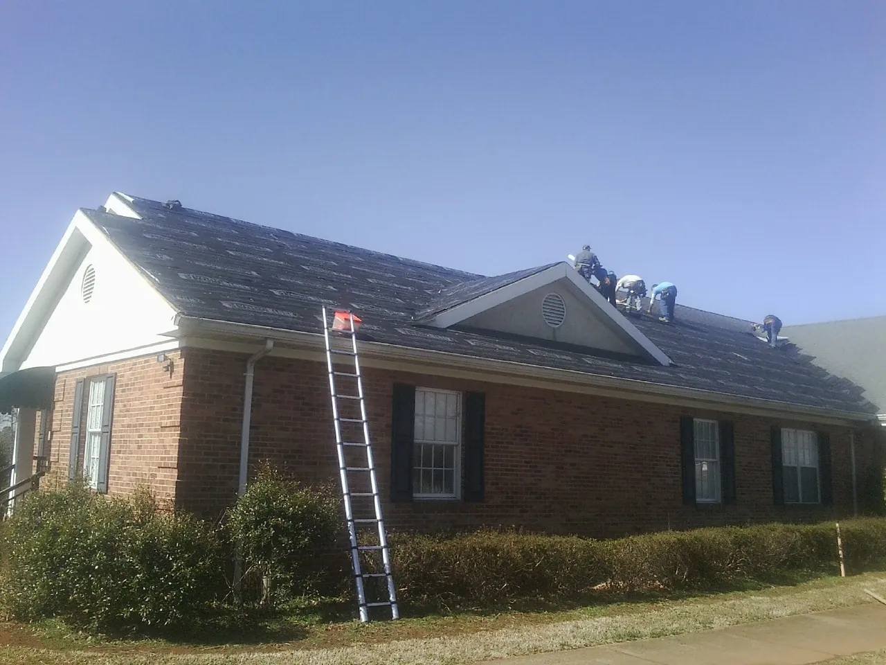 Crew tearing shingles off a brick ranch roof with a ladder set — Emergency Roof Repair work by our crew in Ozone Park
