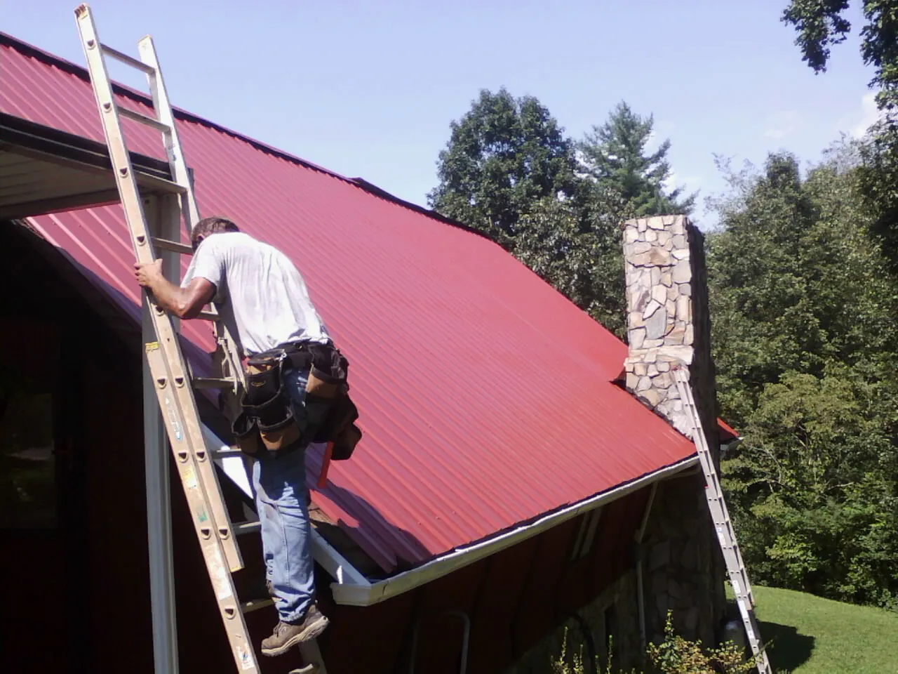 Roofer on a ladder beside a red metal roof and stone chimney — Soffit & Fascia Repair by Roofing Brothers in Ozone Park